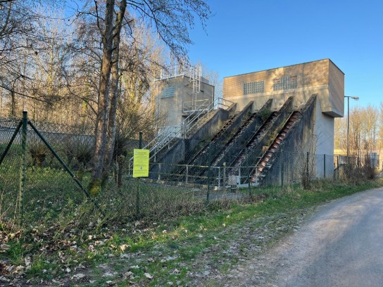 Watergemaal met drie schuine buizen, omgeven door een hek en bomen, onder een blauwe lucht. (vergroot foto)
