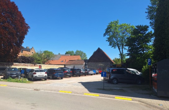Parkeerplaats met meerdere auto's, omgeven door bomen en oude gebouwen onder een heldere blauwe lucht. (vergroot foto)