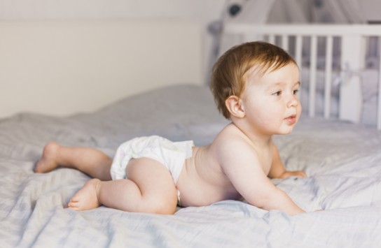 Baby met luier ligt op buik op bed, kijkt naar rechts, op de achtergrond een ledikant. (vergroot foto)