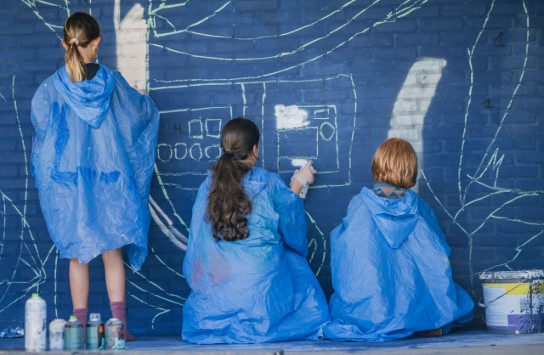 Drie kinderen in blauwe poncho's tekenen met krijt op een blauwe bakstenen muur. (vergroot foto)
