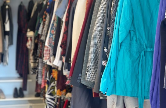 Kledingrekken met diverse jassen, blouses en jurken in een winkel, met opgevouwen kleding op planken eronder. (vergroot foto)