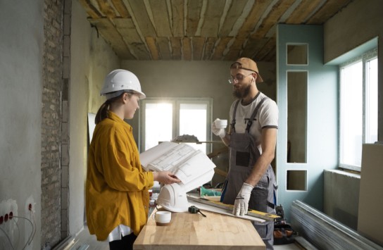 Twee bouwvakkers bespreken bouwplannen in een half afgewerkte kamer met gereedschap op tafel. (vergroot foto)