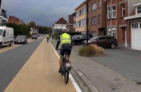 Twee fietsers in gele hesjes fietsen op een apart fietspad langs een woonstraat met geparkeerde auto's. (vergroot foto)