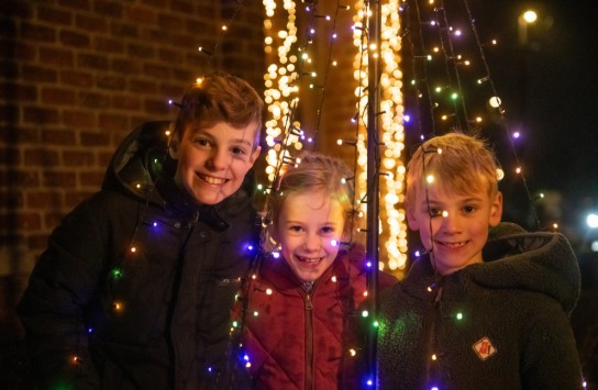 Drie lachende kinderen poseren tussen kleurrijke kerstverlichting. (vergroot foto)
