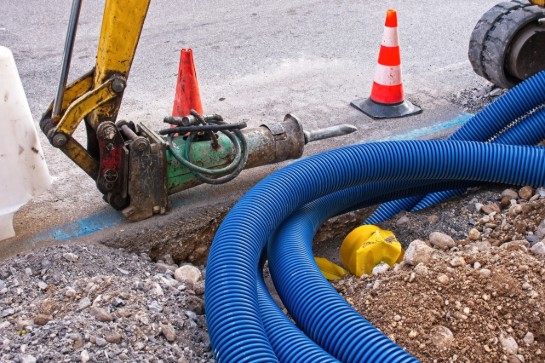 Bouwplaats met graafmachine, blauwe buizen, oranje verkeerskegels en gegraven grond naast asfaltweg. (vergroot foto)