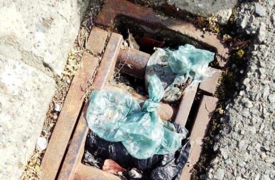 Afval van plastic zakken ligt vast in een roestige rioolgoot tussen asfalt en beton. (vergroot foto)
