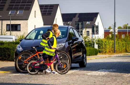 Twee kinderen met fiets en hesjes steken voorzichtig over bij een zwarte auto in een woonwijk met zonnepanelen. (vergroot foto)